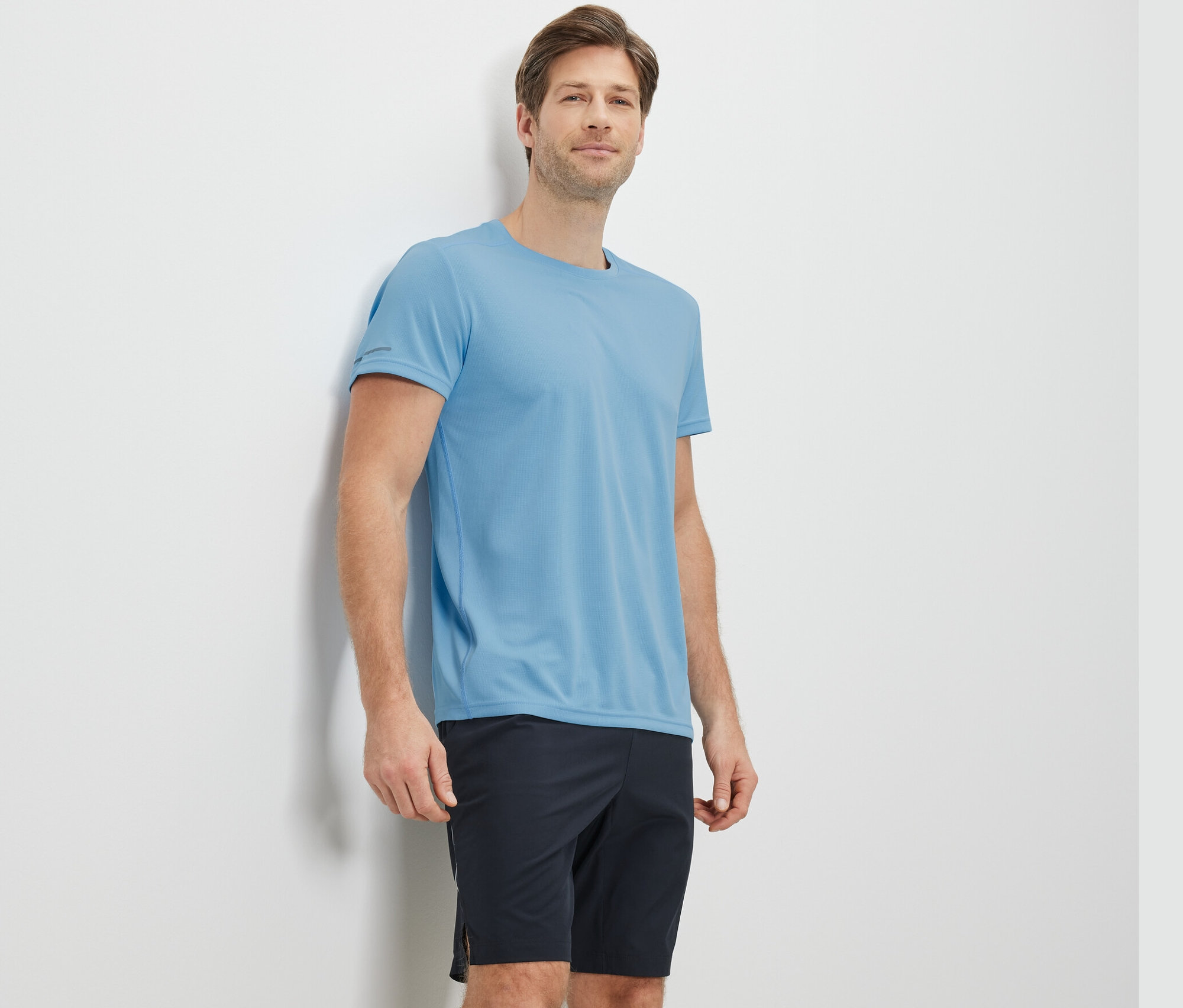 Duvara yaslanmış, açık mavi fonksiyonel tişört giyen adam.