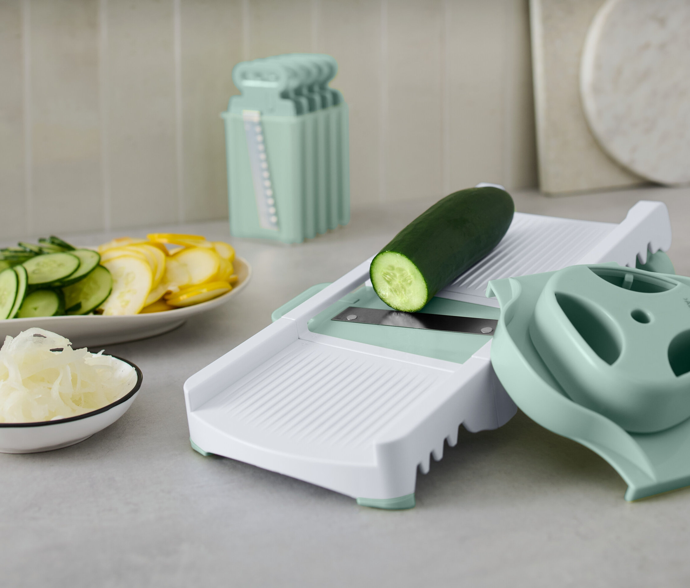 Beyaz bir sebze dilimleyicide aksesuarlarıyla birlikte duran yeşil salatalık. Tabakta dilimlenmiş salatalık ve sarı kabak var.