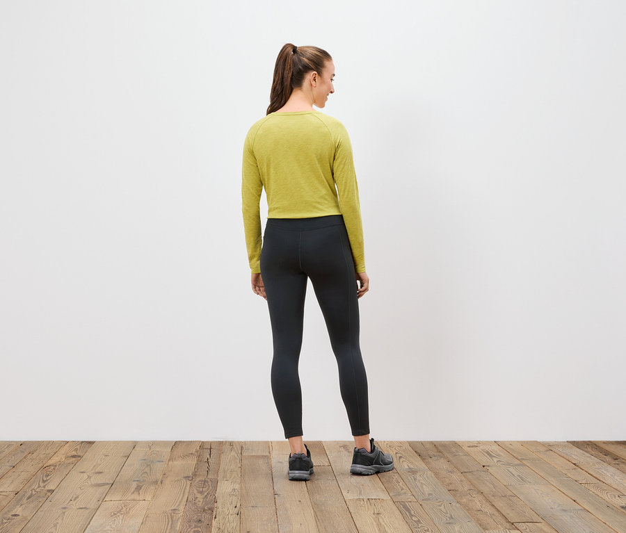 Sırtı kameraya dönük bir kadın, zeytin yeşili uzun kollu bir üst, siyah tayt ve spor ayakkabı giyiyor.