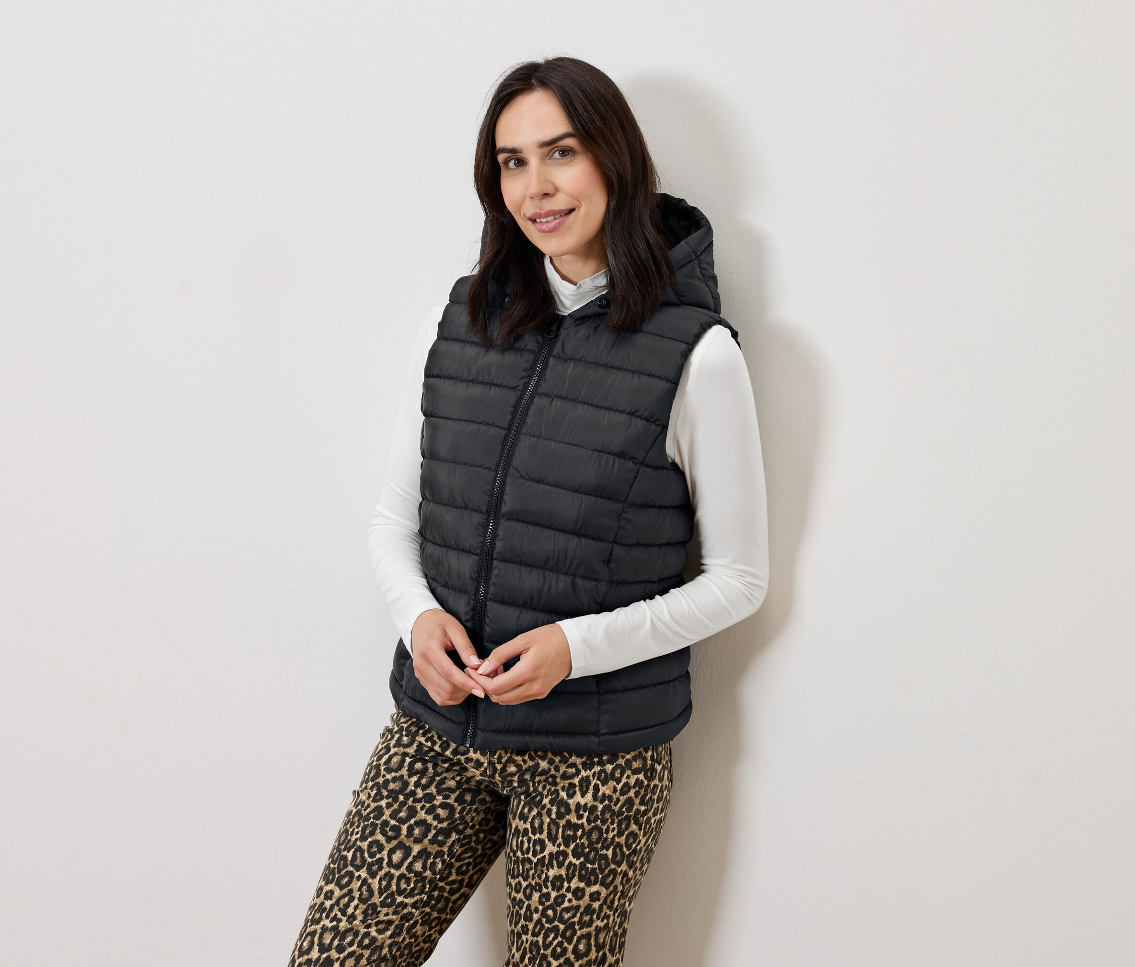 Kadın kapüşonlu siyah kapitone yelek, beyaz balıkçı yaka kazak ve hayvan desenli pantolonla poz veriyor.