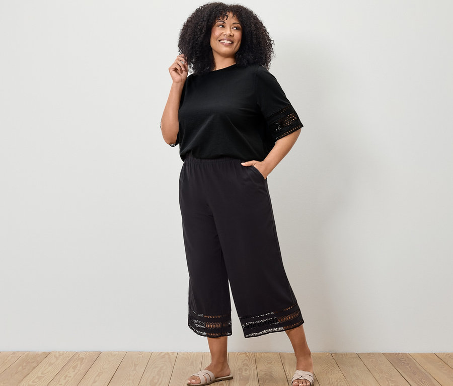 Kadın siyah işlemeli tişört ve siyah işlemeli culotte pantolonla poz veriyor.