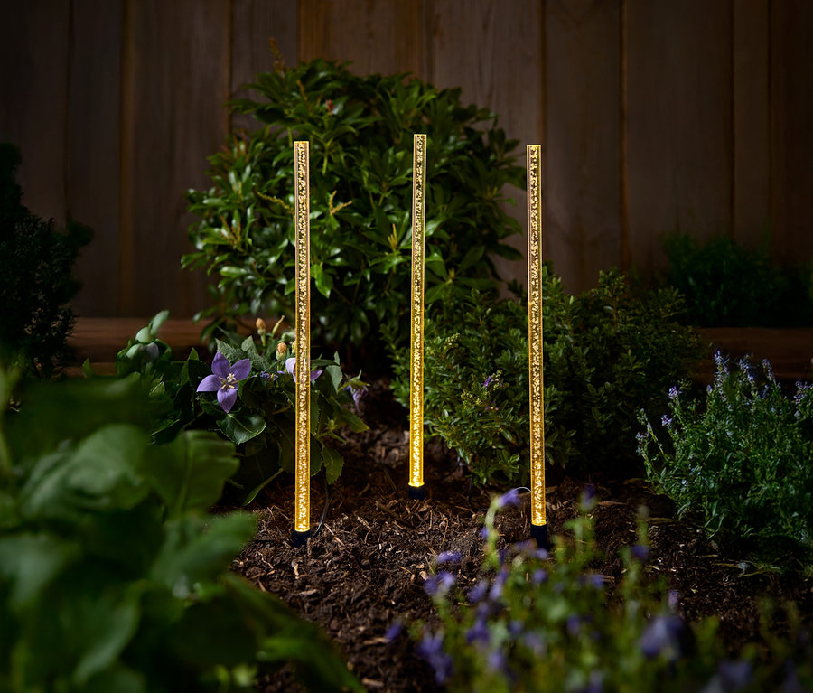 Çalılar ve çiçeklerle dolu bir bahçede duran üç adet altın rengi güneş enerjili bahçe lambası.