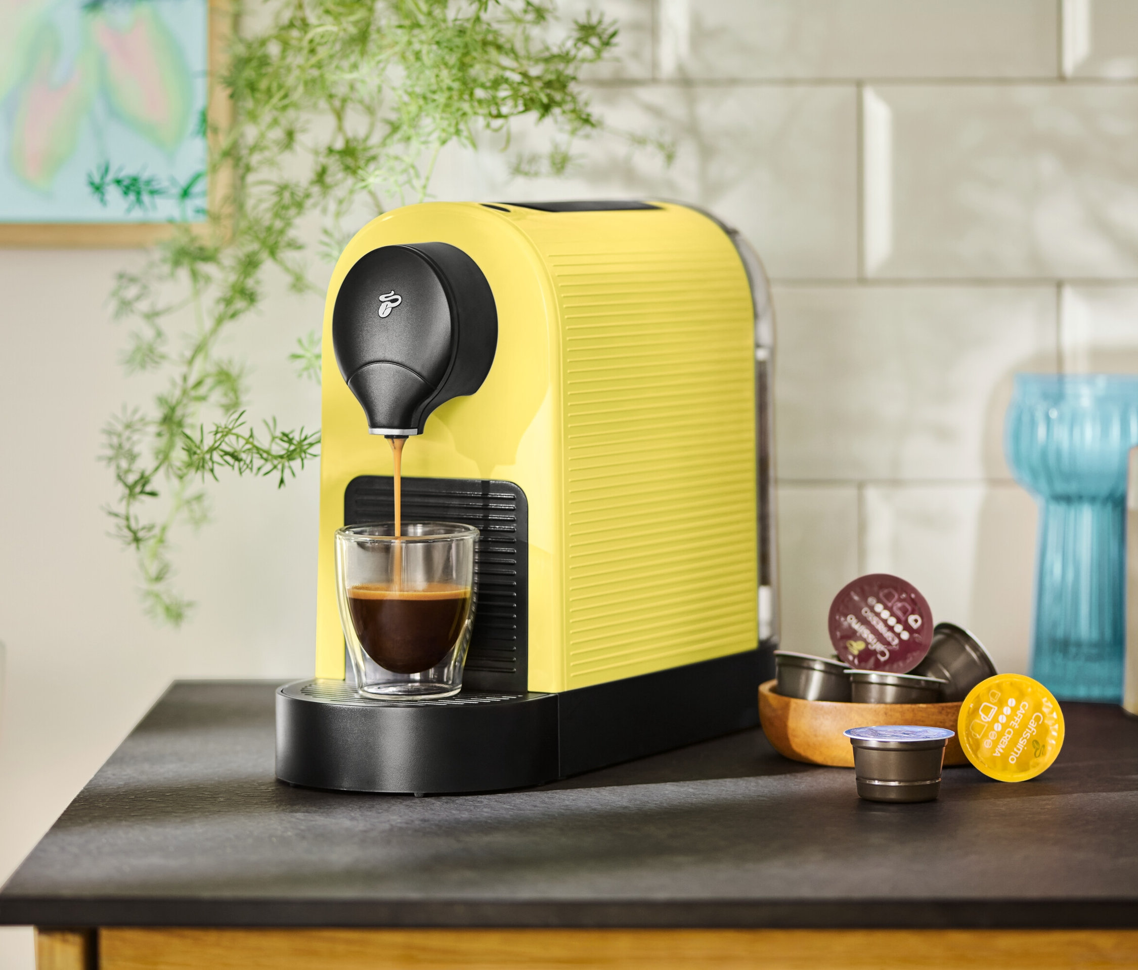 Sarı bir kahve makinesi bir fincanı kahveyle dolduruyor, yanında kapsüller var.