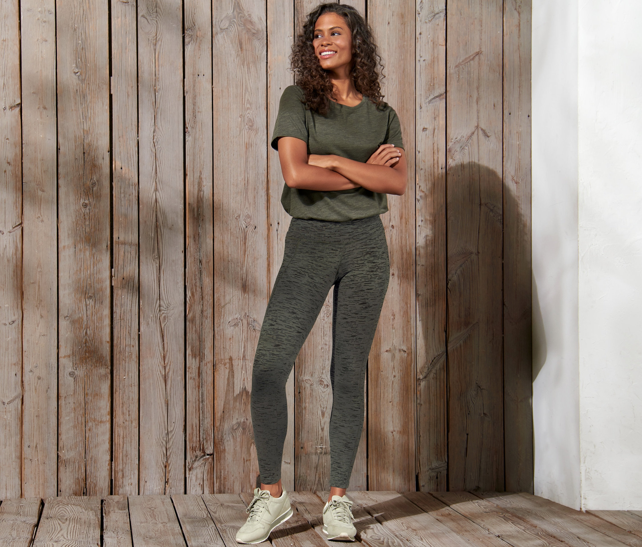 Gülümseyen bir kadın kolları çapraz bir şekilde poz veriyor ve spor taytı giyiyor.