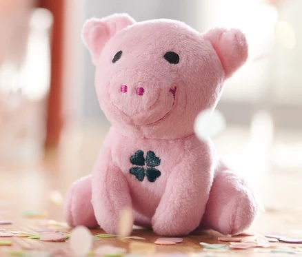 Oturmuş, karnında yonca yaprağı olan, pembe Ses Çıkaran Şans Domuzu, etrafı konfetiyle çevrili.