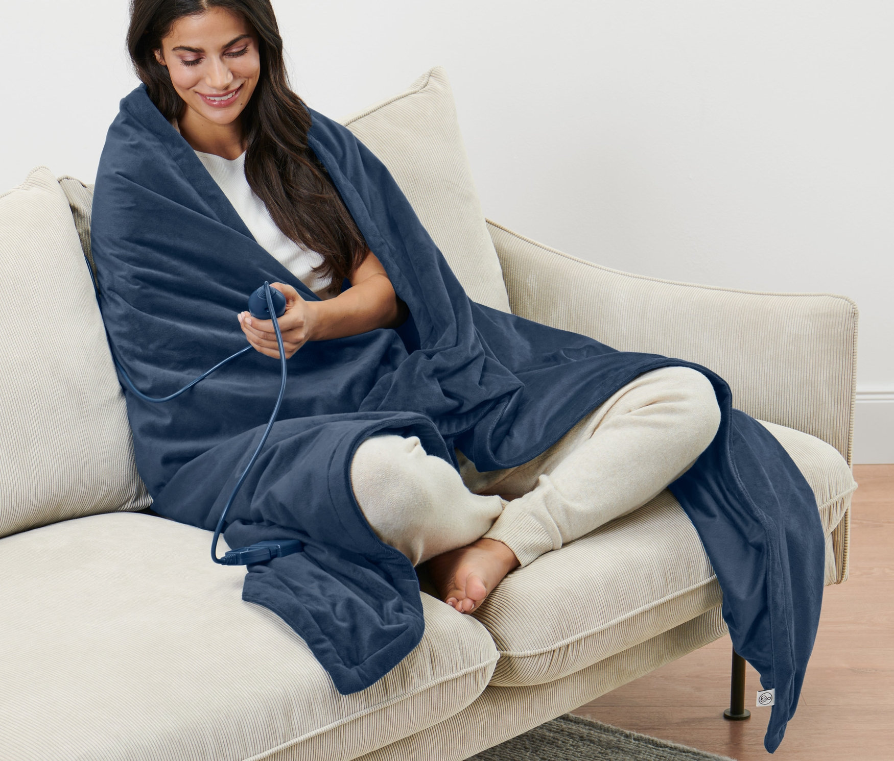 Kadın bej renkli bir kanepede oturuyor, üzerine ısıtıcı battaniye örtmüş.