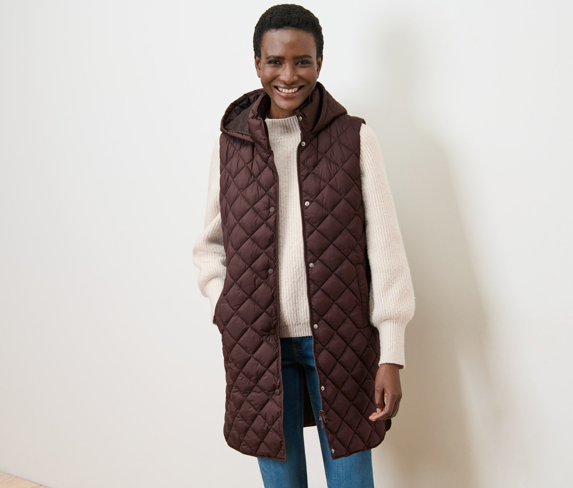 Gülümseyen bir kadın kapüşonlu uzun kapitone yelek ile poz veriyor.