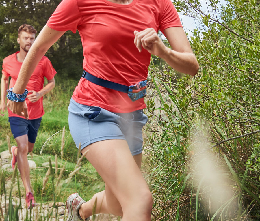 Mercan rengi fonksiyonel tişört ve lacivert 3/4 spor taytlı bir kadın taşlı bir yolda koşuyor. Arka planda fonksiyonel şort ve mercan rengi spor tişörtü giyen bir adam koşuyor.