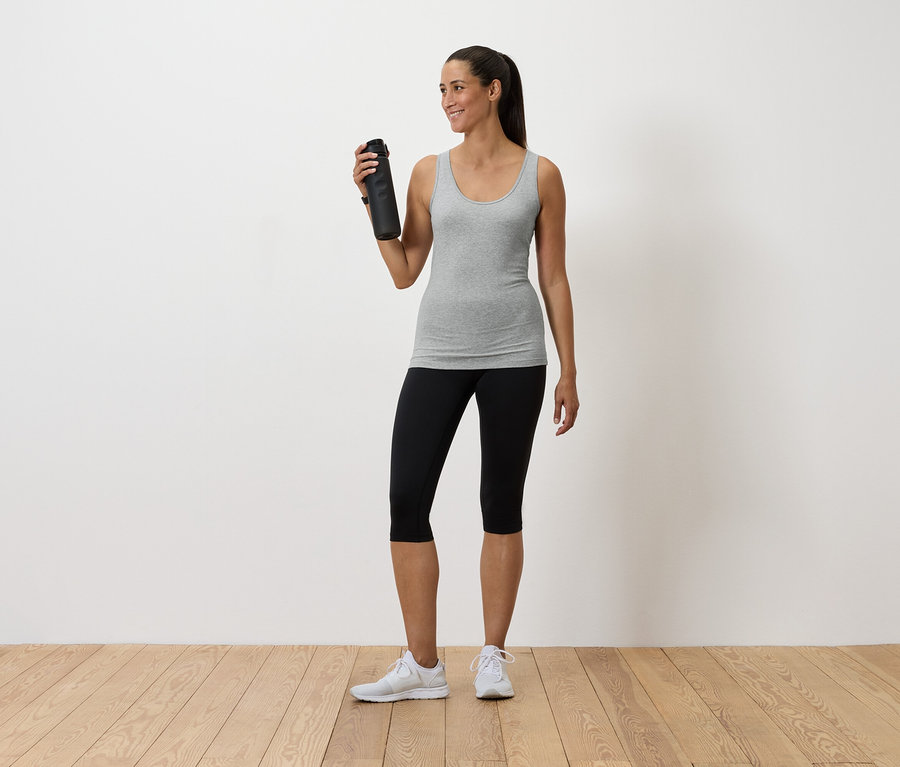 Kadın elinde su şişesi tutuyor, gri atlet, siyah tayt ve beyaz spor ayakkabı giyiyor.