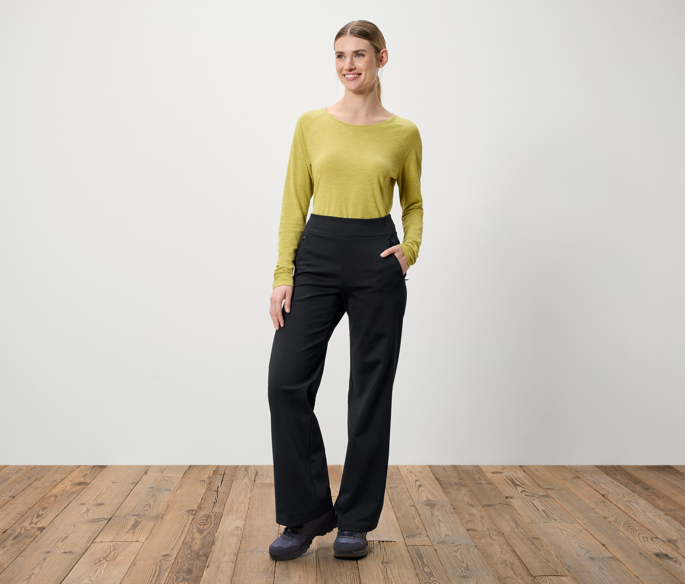 Bir kadın ahşap bir zeminde duruyor. Üzerinde uzun kollu yeşil bir bluz ve siyah pantolon var.