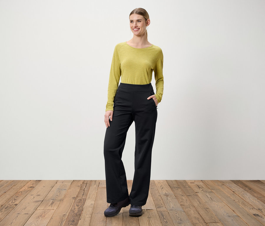 Bir kadın ahşap bir zeminde duruyor. Üzerinde uzun kollu yeşil bir bluz ve siyah pantolon var.