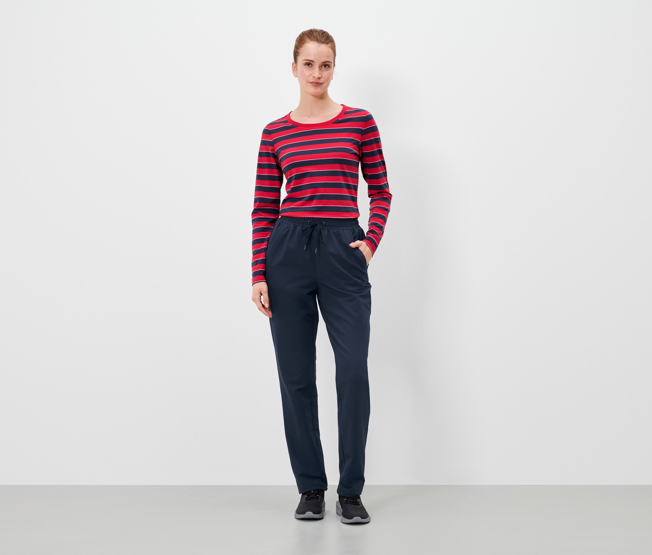 Kadın lacivert pantolonunun ceplerinde elleriyle duruyor ve kırmızı-mavi çizgili bir tişört giyiyor.
