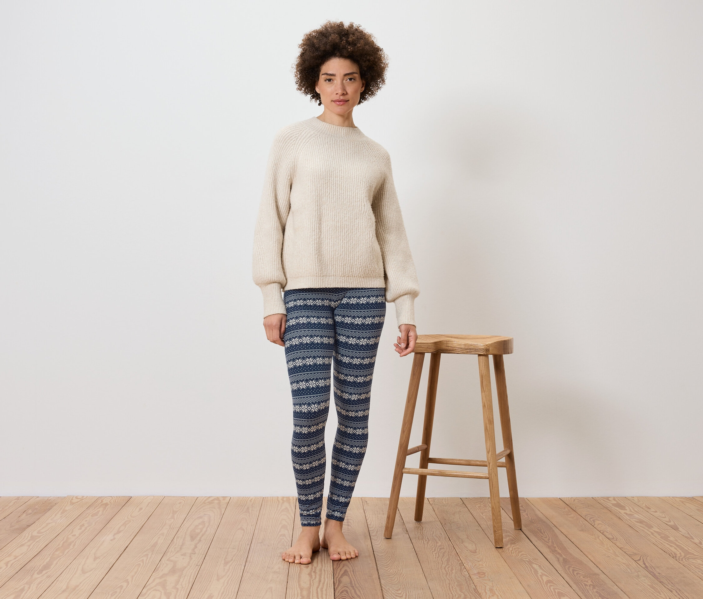 Bir kadın beyaz bir duvarın önünde, ahşap bir taburenin yanında duruyor. Üzerinde krem rengi bir kazak ve desenli mavi bir tayt var.