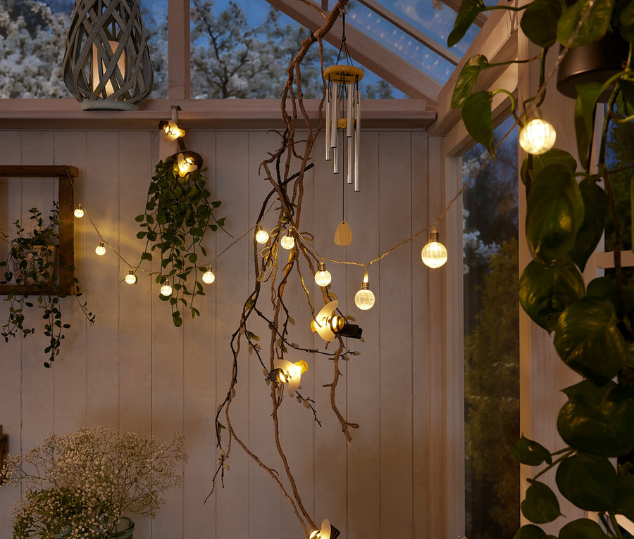 Gri bir fener, küre ve arı desenli ışık zinciri, rüzgar çanı ve bitkilerin bulunduğu bir kış bahçesinin iç mekanı.