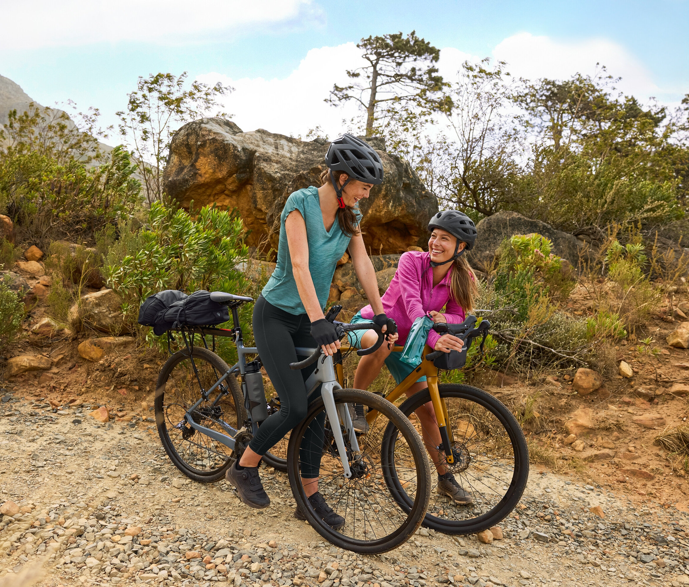 Kasklı iki kadın, engebeli arazide çakıllı bir yolda bisikletleriyle duruyor. Bir matara bisiklete takılı.