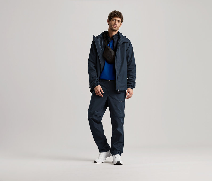 Lacivert ceket, pantolon ve beyaz spor ayakkabı giyen, siyah bir çapraz çantası olan bir adam ayakta duruyor.