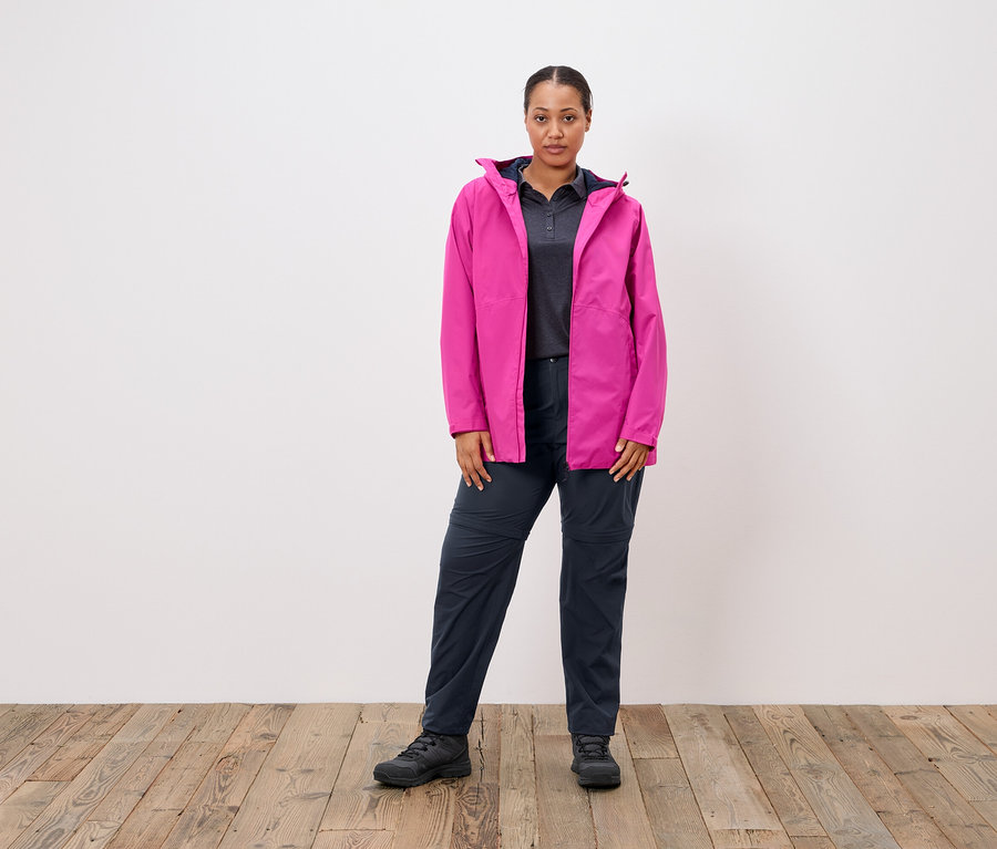 Kadın beyaz bir duvarın önünde duruyor, pembe bir ceket, gri bir polo tişört ve lacivert pantolon giyiyor.