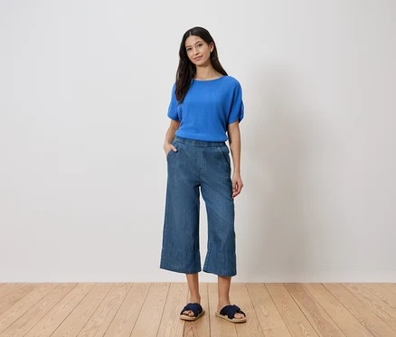 Kadın eli cebinde duruyor, beyaz bir duvarın önünde mavi bir bluz, denim culottes ve sandalet giyiyor.