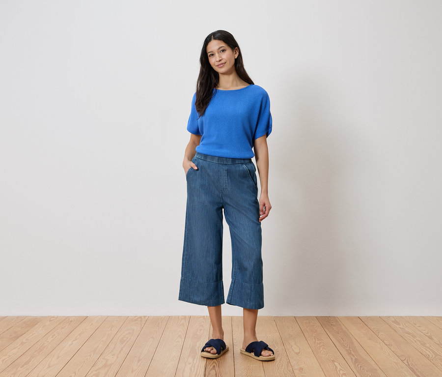 Kadın eli cebinde duruyor, beyaz bir duvarın önünde mavi bir bluz, denim culottes ve sandalet giyiyor.
