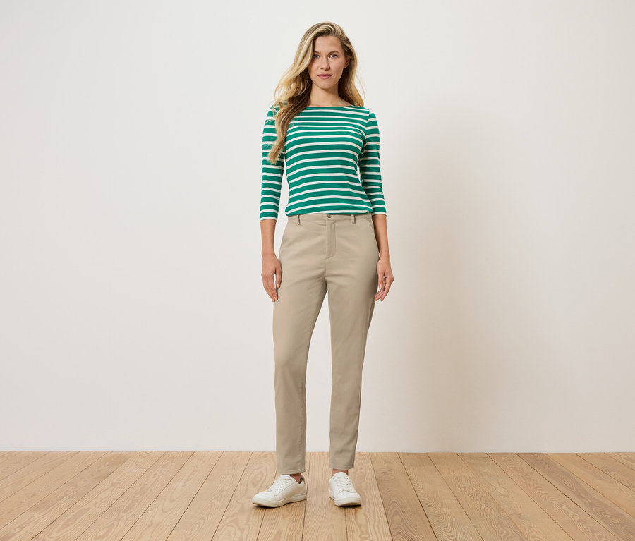 Kadın ayakta duruyor, yeşil ve beyaz çizgili uzun kollu bir bluz, bej pantolon ve beyaz ayakkabılar giyiyor.