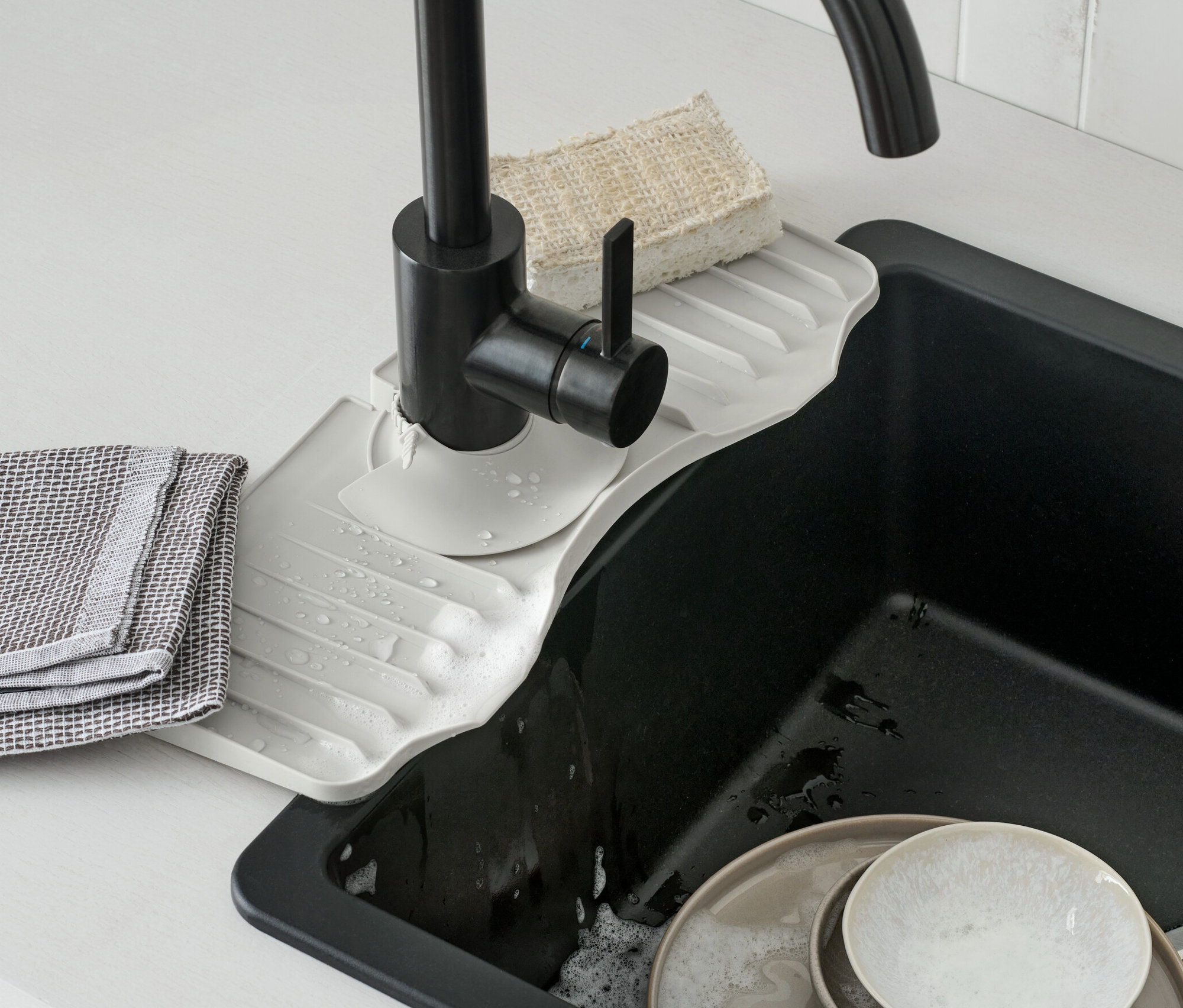 Silikon Musluk Rafı siyah bir musluğun altına yerleştirilmiştir. Lavabonun içinde kirli bulaşıklar var.