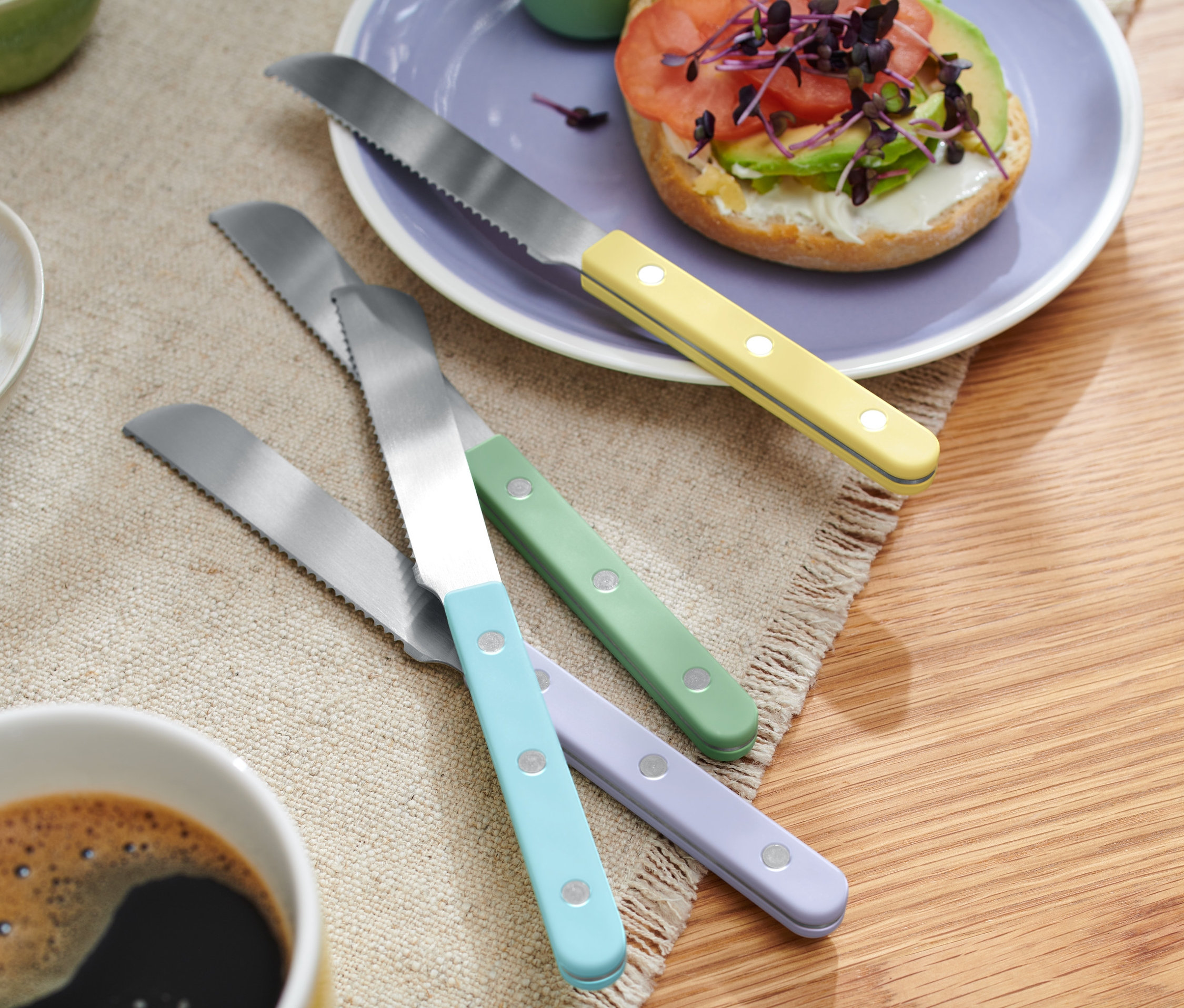 Dört renkli kahvaltı bıçağı, bir simit tabağı ve bir fincan kahvenin yanında bir masanın üzerinde duruyor.