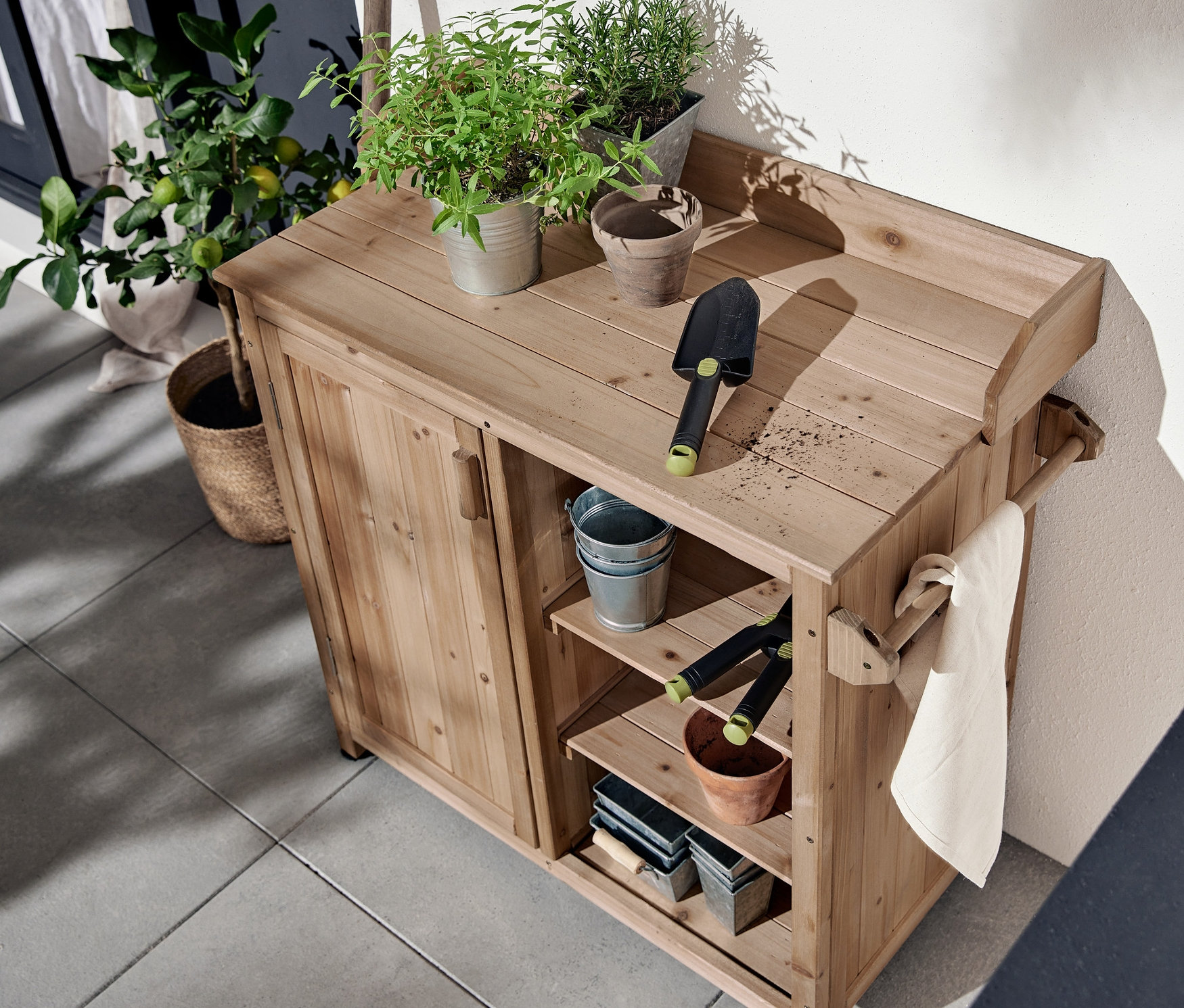 Verandada çiçeklerin ve bahçe aletlerinin üzerinde bulunduğu havlu askılı bahçe dolabı duruyor.