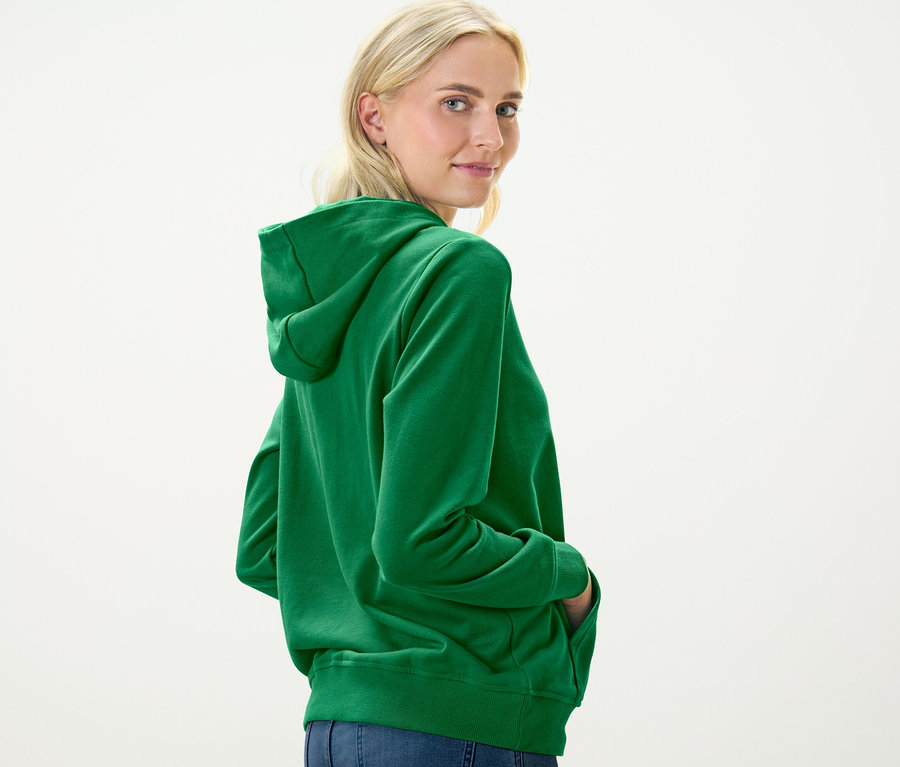 Sarışın bir kadın omzunun üzerinden bakıyor ve yeşil bir kapüşonlu sweatshirt giyiyor.