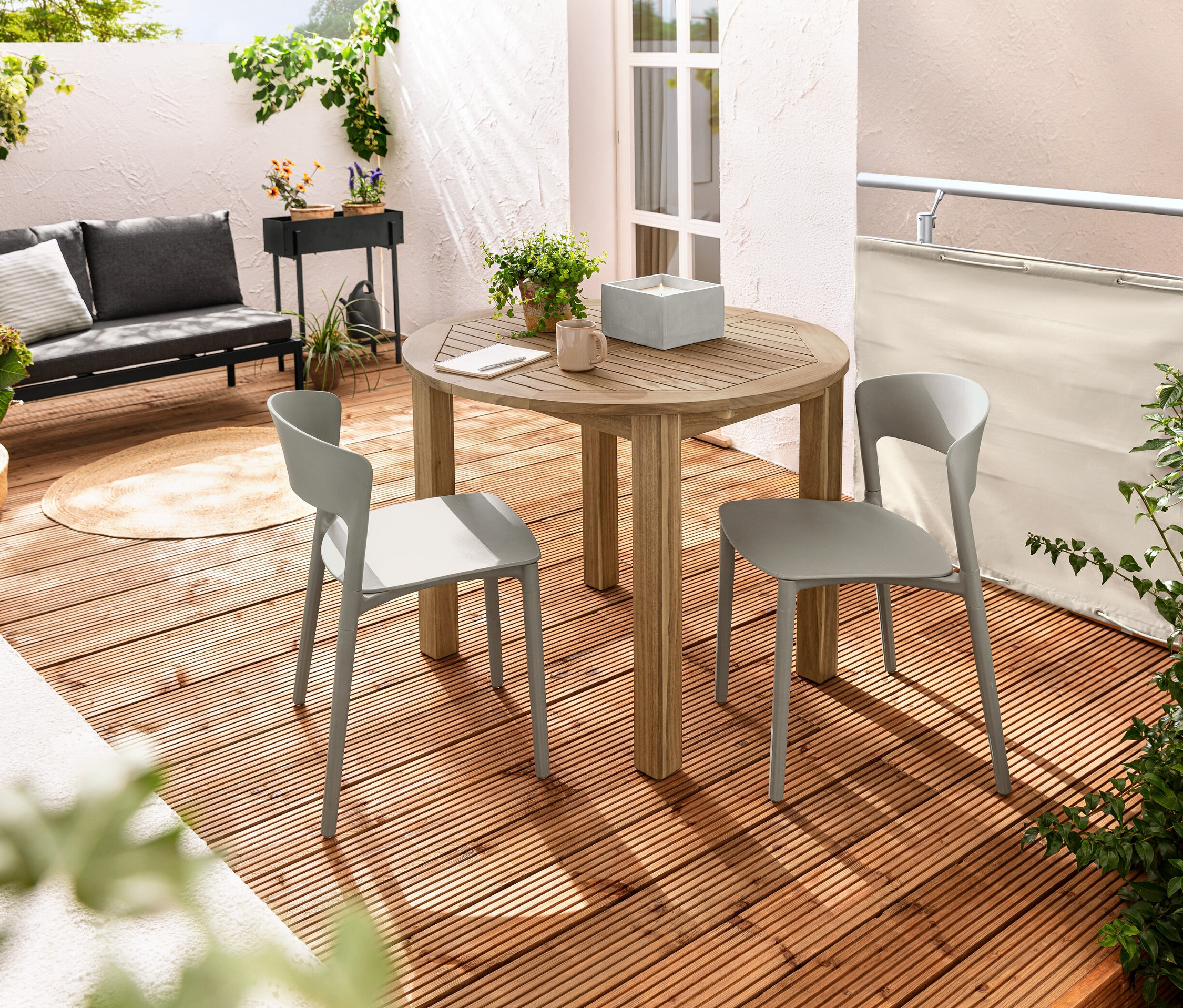 Balkonda yuvarlak açılır masanın yanında iki adet plastik sandalye duruyor. Masanın üzerinde beton saksıda XL LED gerçek balmumu mum var. Arka planda krem rengi balkon paravanı ve rüzgar koruması görülüyor.