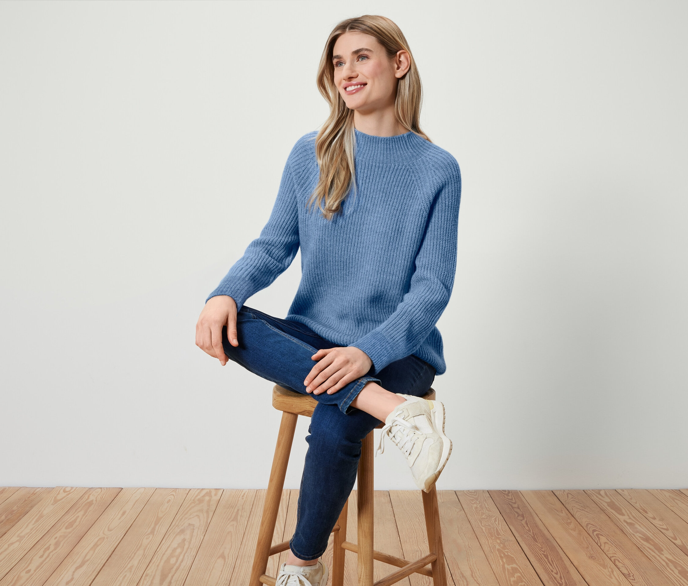 Kadın ahşap bir taburede oturuyor, mavi bir kazak, koyu mavi kot pantolon ve beyaz spor ayakkabısı giyiyor.