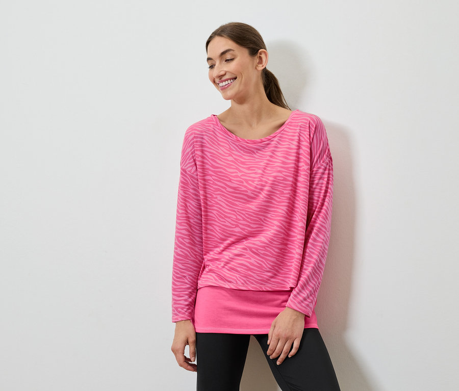 Gülümseyen bir kadın pembe uzun kollu bir üst ve siyah taytla poz veriyor.