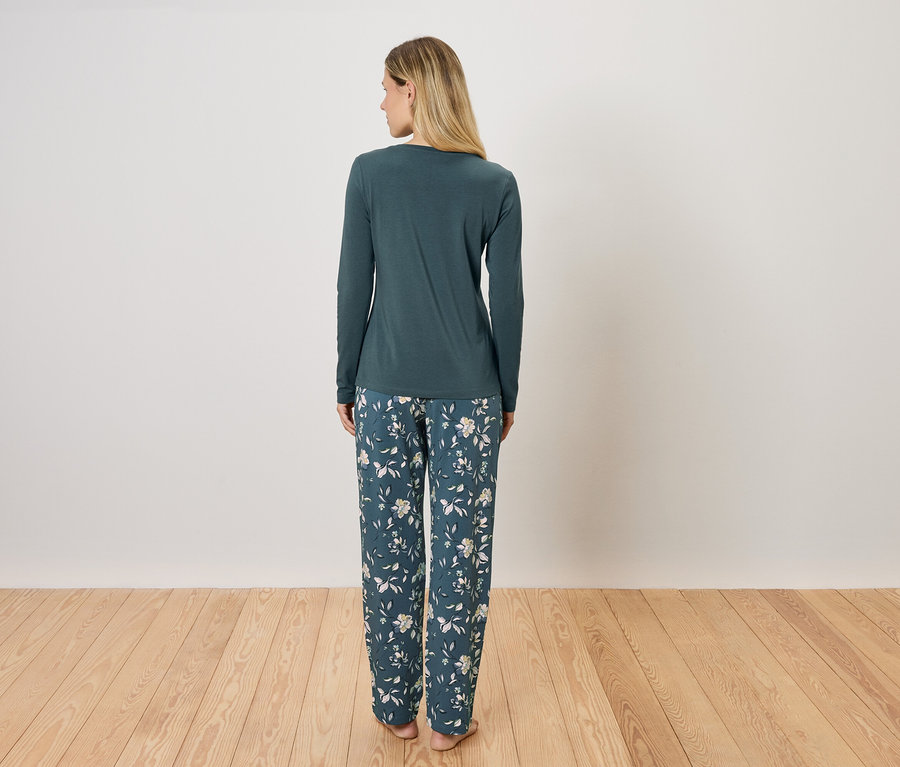 Kadın arkası dönük duruyor, üzerinde uzun kollu yeşil bir pijama ve çiçekli bir pantolon var.