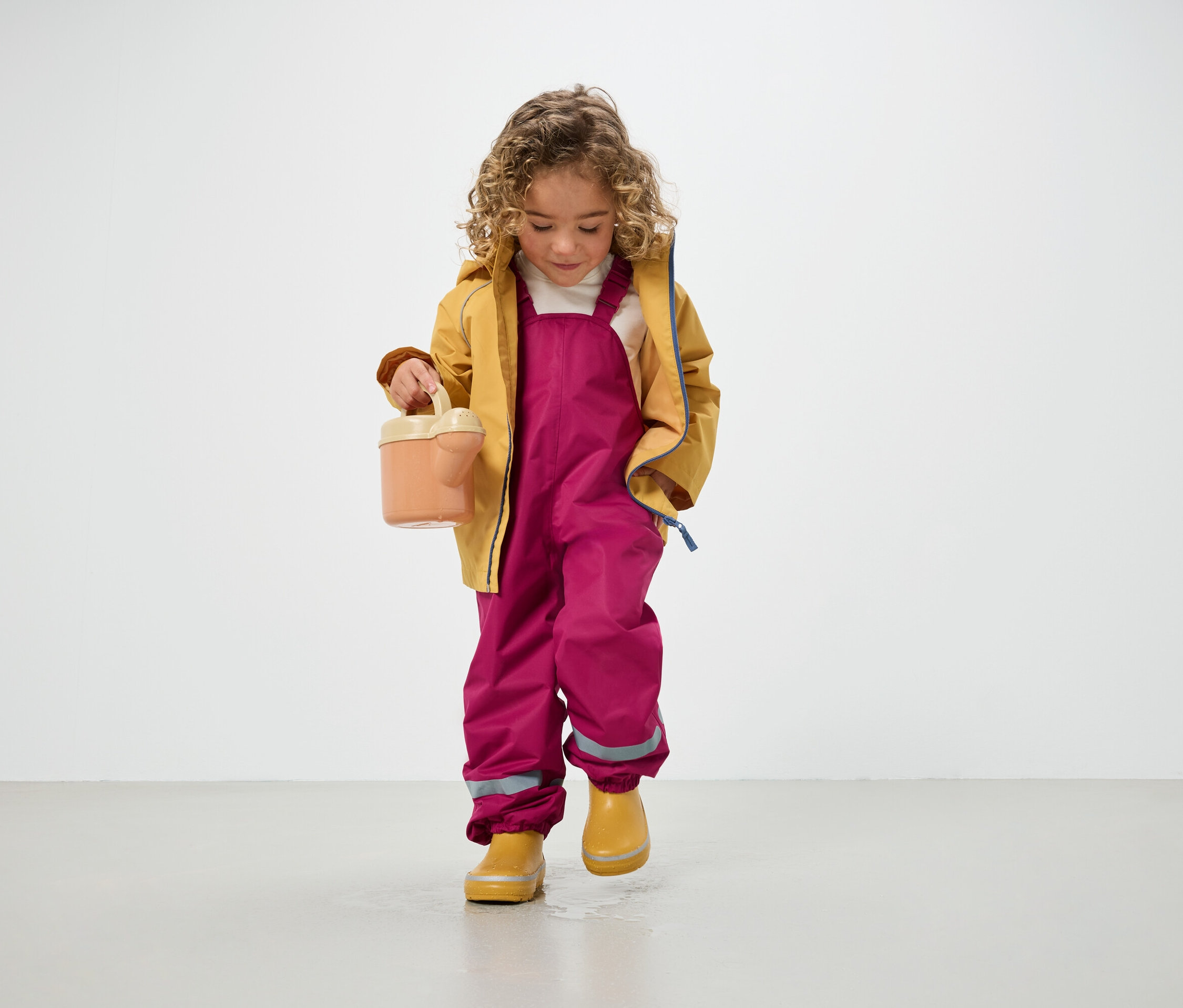 Sarışın kıvırcık saçlı çocuk sarı mont, pembe pantolon ve sarı çizme giyiyor, elinde sulama kabı tutuyor ve yerde duruyor.