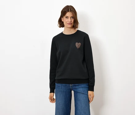 Kadın ayakta duruyor, üzerinde kalp şeklinde süsü olan siyah bir sweatshirt ve kot pantolon var.