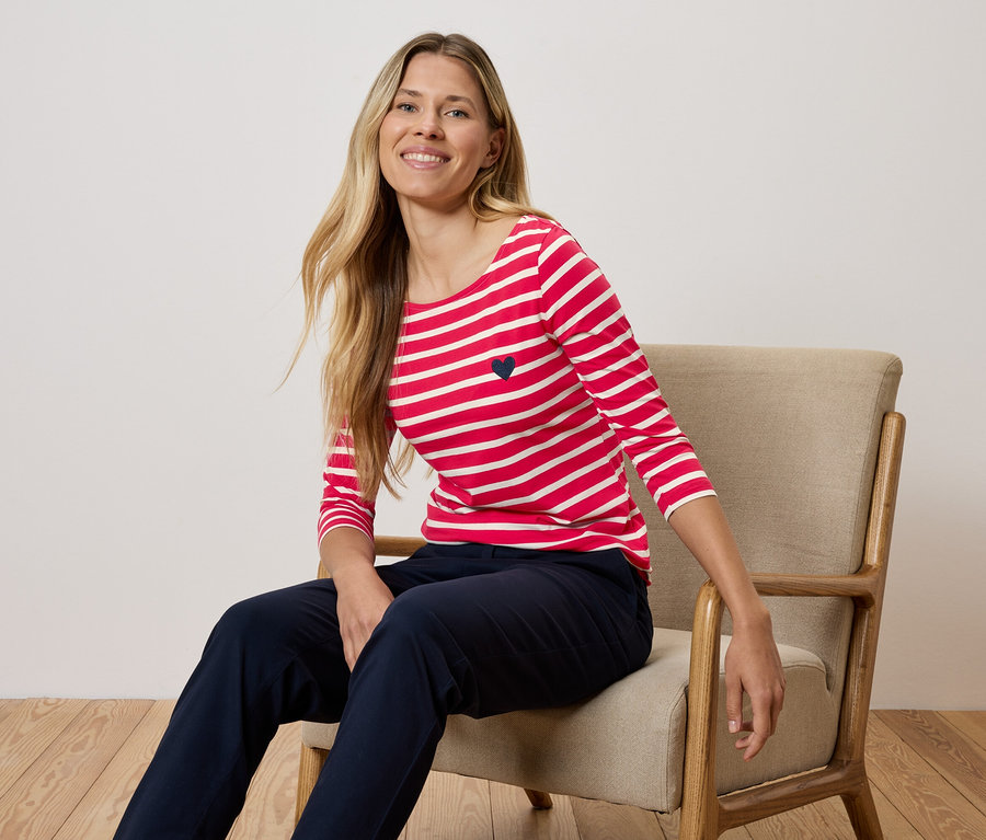 Bir kadın sandalyede oturuyor, kırmızı beyaz çizgili tişört ve lacivert pantolon giyiyor.