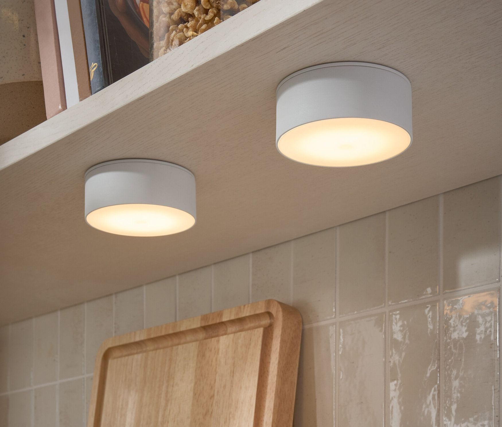 Bir rafın altına takılmış iki adet LED çok fonksiyonlu lamba. Rafta fındık kavanozları ve kitaplar var.