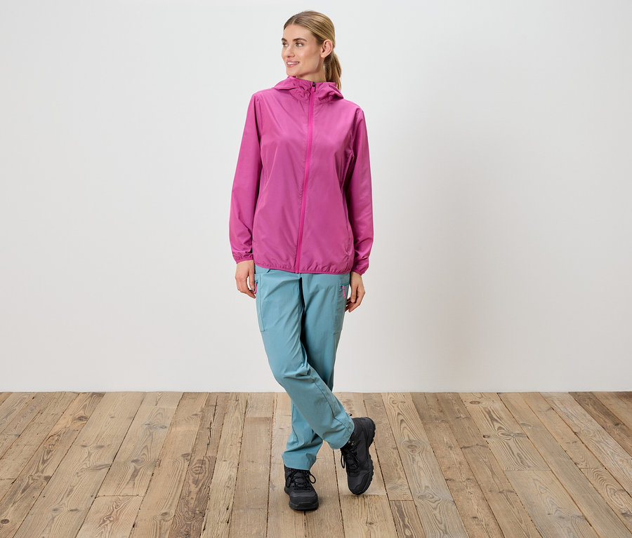 Kadın çapraz bacaklı duruyor, pembe ceket, mavi pantolon ve siyah ayakkabılar giyiyor.