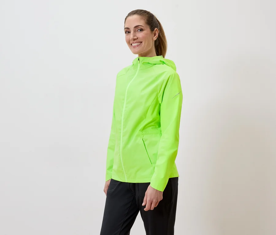 Atkuyruklu kadın beyaz bir arka planda neon yeşili bir yağmurluk ve siyah pantolon giyiyor.