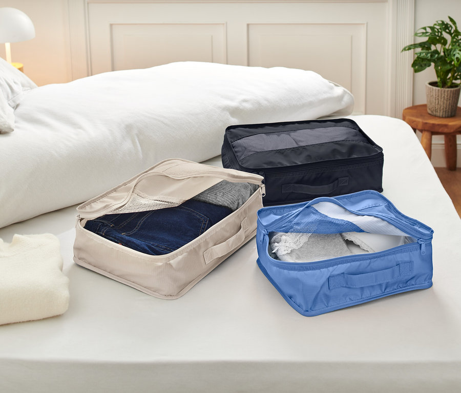 Beyaz çarşaflı bir yatak üzerinde üç adet açık bavul düzenleyici bulunmaktadır. Bavul düzenleyiciler bej, mavi ve siyah renktedir ve içlerinde giysiler bulunmaktadır.