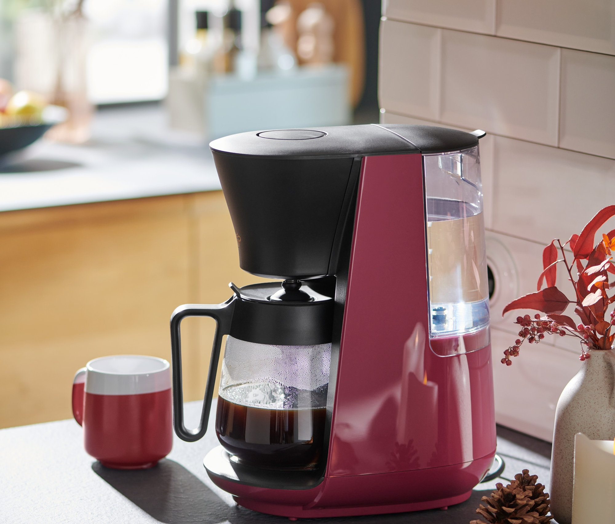 Berry renkli »Let's Brew« filtre kahve makinesi, mutfak tezgahında kırmızı bir kahve fincanının yanında duruyor.