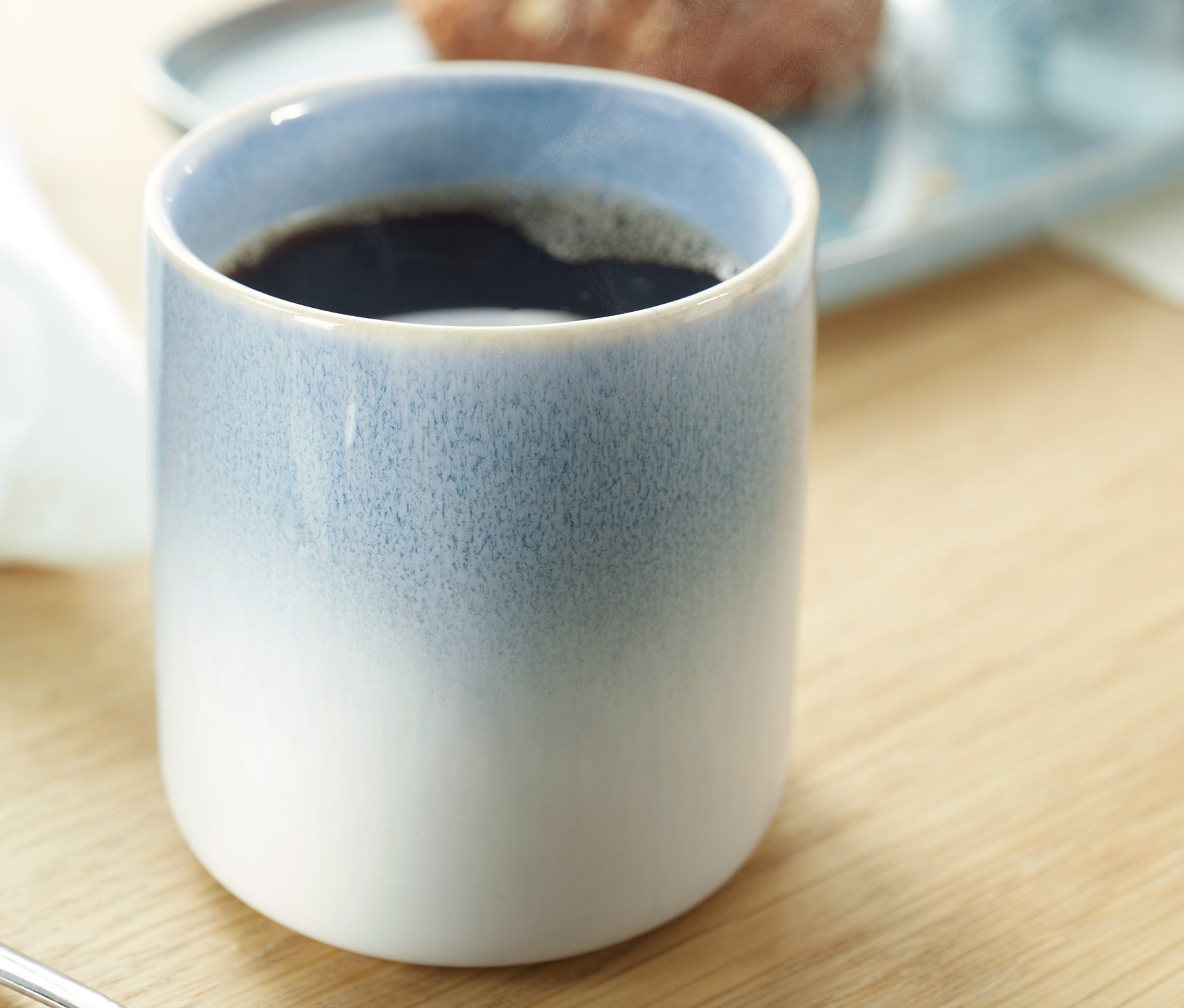 Kahve kupasının yakın çekimi. Arka planda yemek olan bir tabak görülüyor.