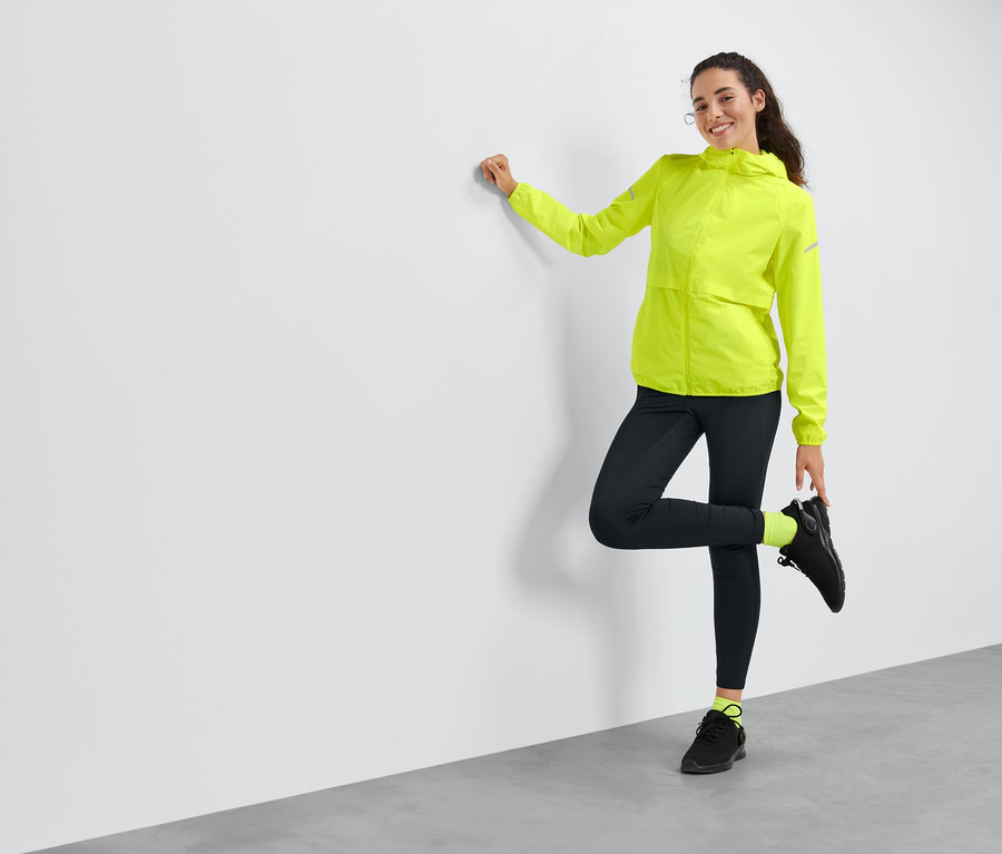 Duvara yaslanmış, sarı rüzgar korumalı koşu ceketi giyen bir kadın poz veriyor.