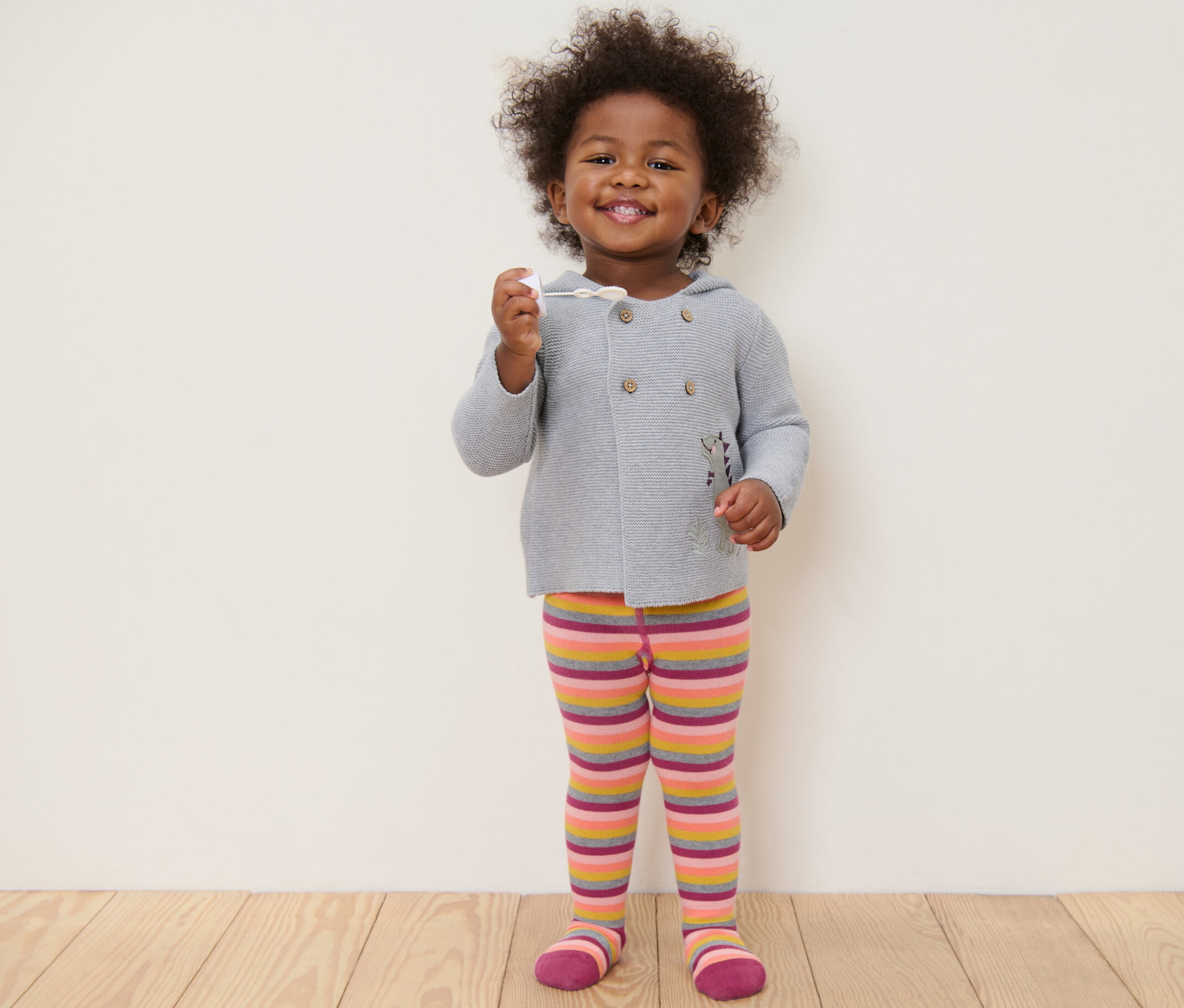 Gülümseyen bir çocuk ayakta duruyor ve elinde bir oyuncak tutuyor. Gri bir kazak ve pembe çizgili külotlu çorap giyiyor.