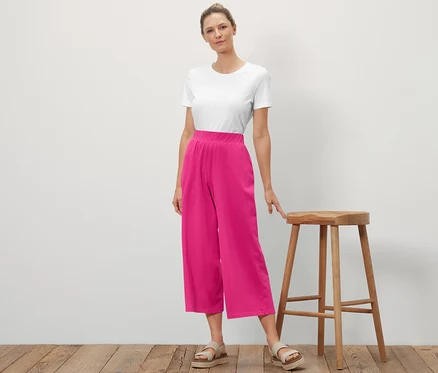 Bir kadın ahşap bir taburenin yanında duruyor ve poz veriyor. Üzerinde beyaz bir tişört ve pembe renkli Jersey Culotte Pantolon var.