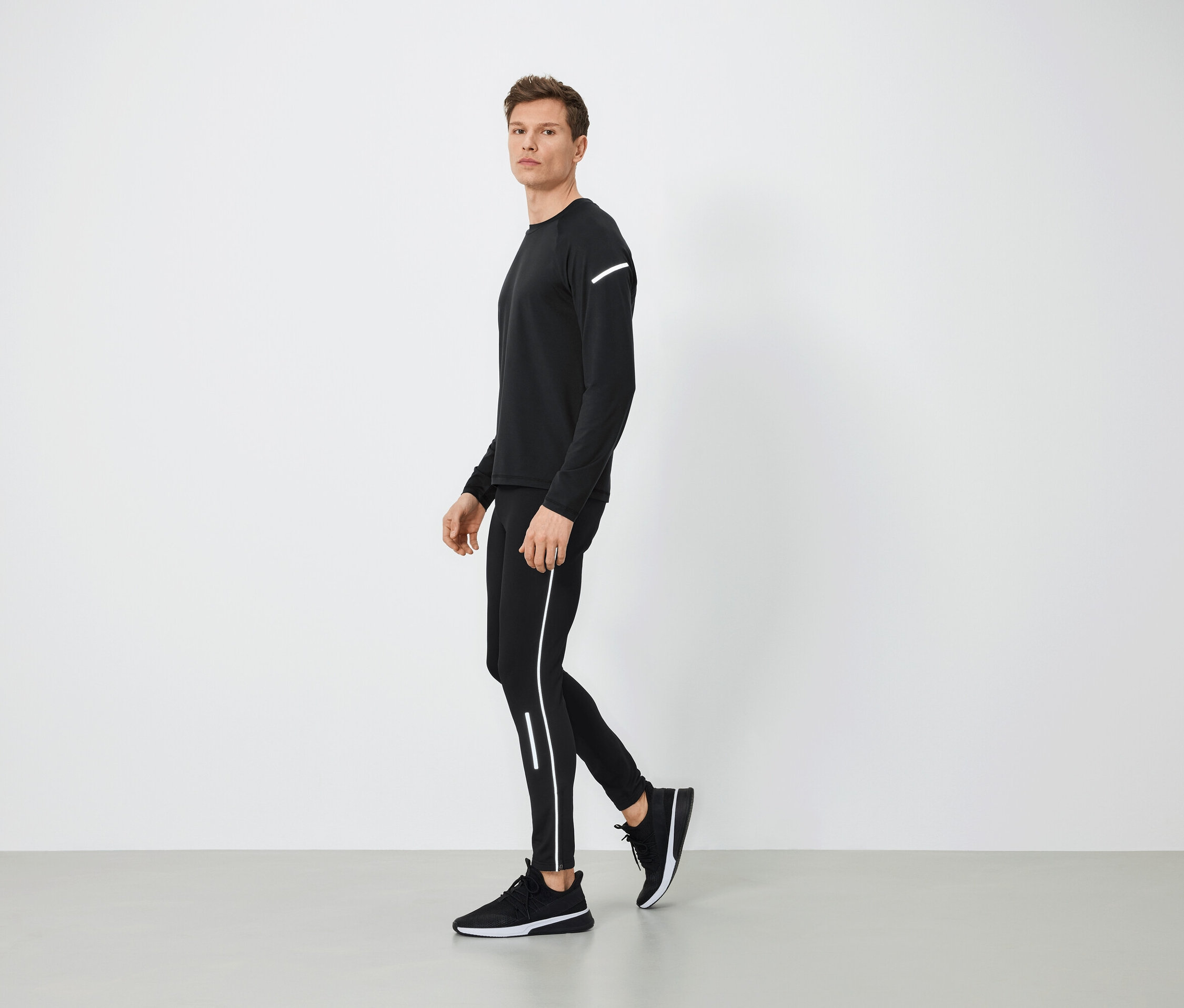 Siyah spor kıyafetli, reflektörlü detaylara sahip bir adam gri zeminde yürüyor.