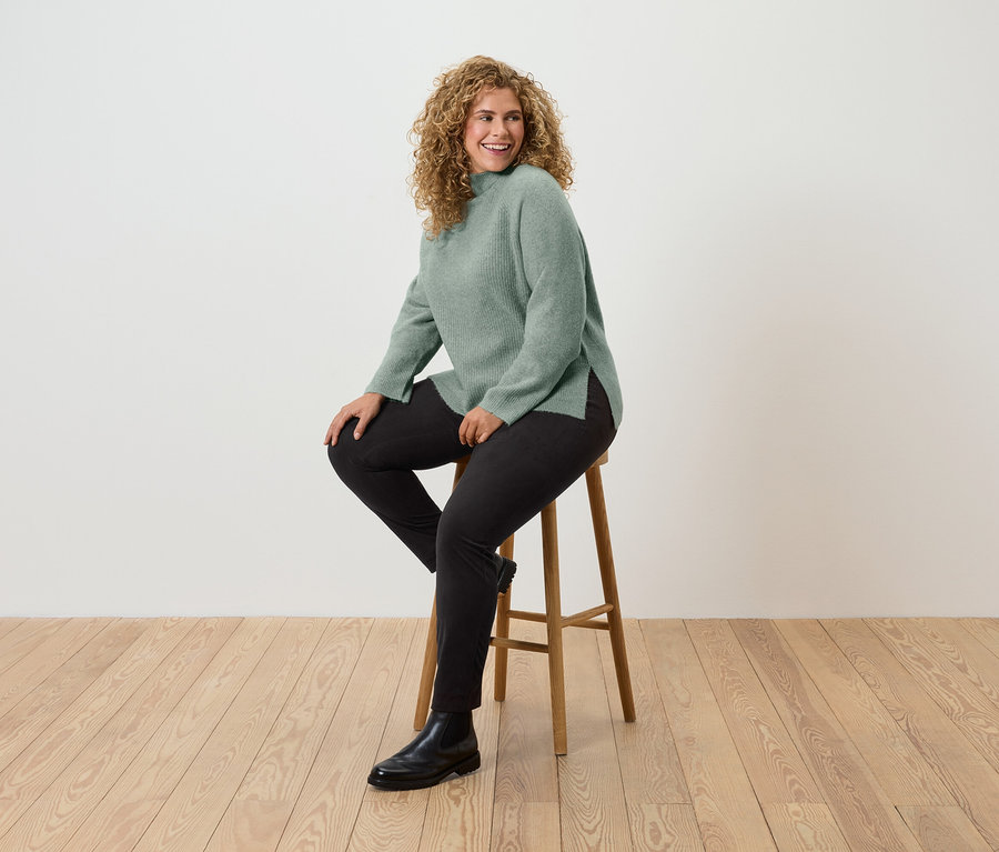 Kıvırcık saçlı bir kadın ahşap bir taburede oturuyor, üzerinde yeşil bir kazak, siyah pantolon ve siyah botlar var.