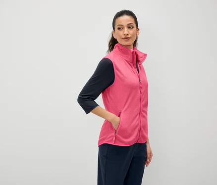 Kahverengi saçlı bir kadın, pembe bir yeleğin cebinde elini tutuyor. Lacivert renkli üç çeyrek kollu bir tişört ve lacivert pantolon giyiyor.