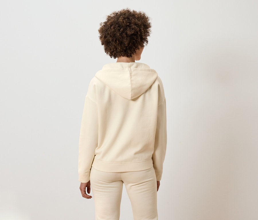 Arkadan kıvırcık saçlı, bej kapüşonlu bir takım elbise giyen bir kadın görünüyor.