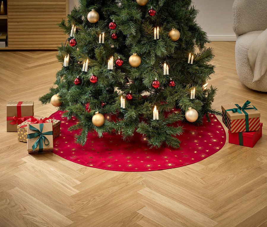 Kırmızı ve altın süslemelerle ve ışıklarla süslenmiş bir Noel ağacı, altın yıldızlarla çevrili kırmızı bir eteğin üzerinde duruyor, etrafı hediyelerle çevrili.