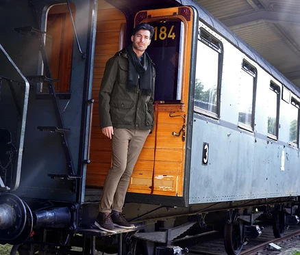 Adam tarihi bir trenin basamaklarında duruyor. Bej rengi bir chino pantolon, yeşil bir ceket ve bir eşarp giyiyor.